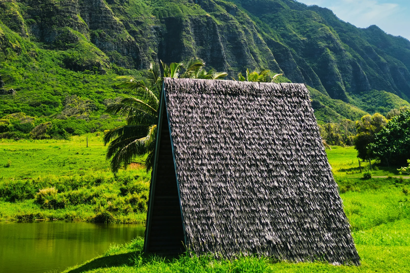 A thatched hale beside a pond, with sunlit green mountain ridges at Kualoa on Oʻahu